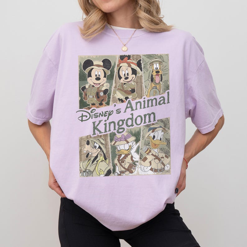 May include: A light purple t-shirt featuring a graphic of Disney's Animal Kingdom characters. The design includes Mickey Mouse, Minnie Mouse, Pluto, Goofy, and Donald Duck in safari outfits. The text "Disney's Animal Kingdom" is also displayed.