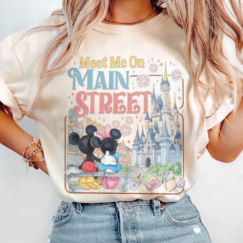 Op de afbeelding: Cr&egrave;mekleurige sweatshirt met een afbeelding van Mickey en Minnie Mouse die elkaar omarmen op een bankje voor een kasteel. De tekst "Meet Me On Main Street" staat in blauw, roze en geel boven de afbeelding.