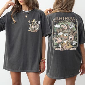 Puede incluir: Camiseta gris oscuro con un dise&ntilde;o de bolsillo con personajes de Disney. La parte trasera de la camiseta muestra un gr&aacute;fico m&aacute;s grande con el texto "ANIMAL KINGDOM" y "Walt Disney World". La camiseta tiene mangas cortas y un corte holgado.