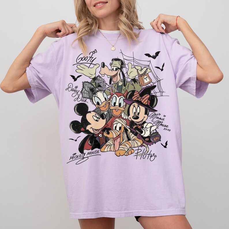 Może przedstawiać: Jasnofioletowy t-shirt z nadrukiem graficznym przedstawiającym postacie Disneya w kostiumach na Halloween, w tym Myszkę Miki, Myszkę Minnie, Goofy'ego, Kaczora Donalda, Daisy i Pluto, z pajęczynami, nietoperzami oraz imionami zapisanymi kursywą.
