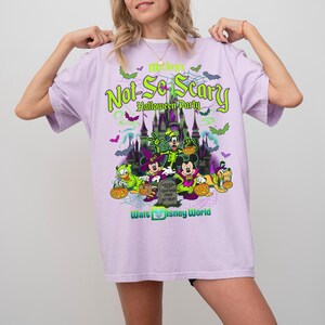 May include: A light purple t-shirt featuring a graphic print of Mickey's Not-So-Scary Halloween Party at Walt Disney World. The design includes a castle, bats, spiderwebs, and characters dressed in costumes holding pumpkins and a tombstone.