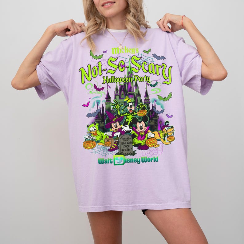 May include: A light purple t-shirt featuring a graphic print of Mickey's Not-So-Scary Halloween Party at Walt Disney World. The design includes a castle, bats, spiderwebs, and characters dressed in costumes holding pumpkins and a tombstone.