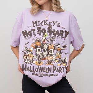 May include: A light purple t-shirt featuring a graphic of Disney characters in skeleton costumes. The text "Mickey's Not So Scary Halloween Party" and "Walt Disney World" are printed on the shirt.