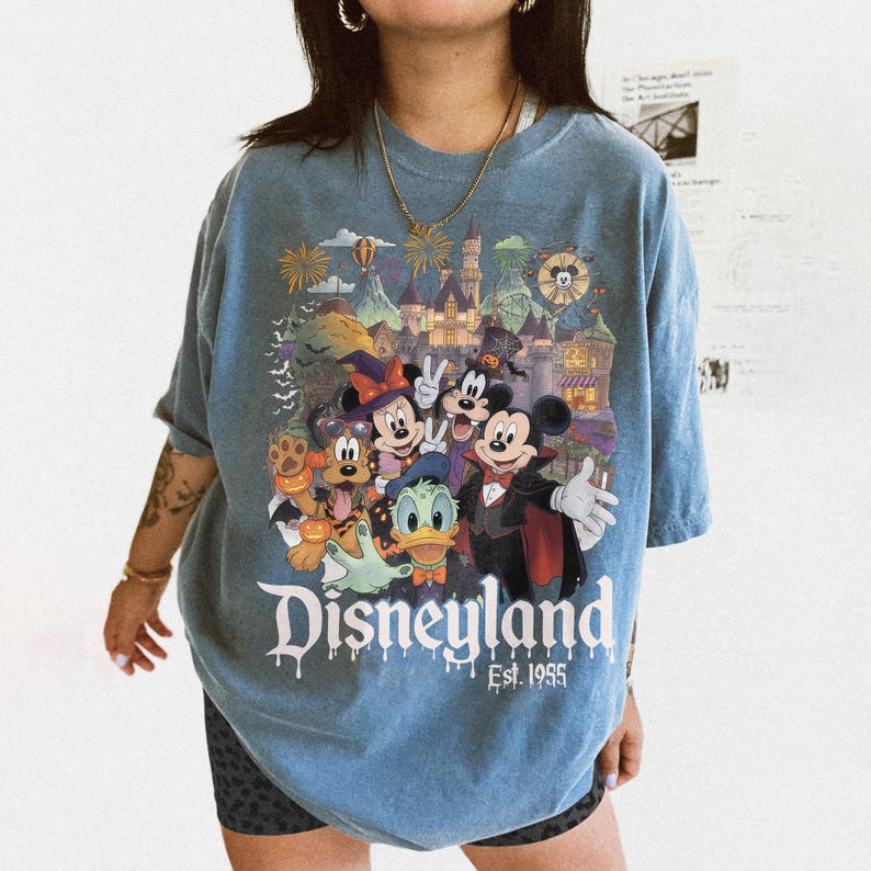 May include: A blue short-sleeve t-shirt featuring a graphic print of Disney characters in Halloween costumes, including Mickey Mouse, Minnie Mouse, Goofy, Donald Duck, and Pluto, with a castle background and the text Disneyland Est. 1955.