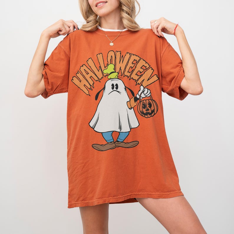 May include: An orange t-shirt with the word "HALLOWEEN" in orange letters. The shirt features a cartoon ghost holding a jack-o'-lantern. The ghost is wearing a green hat, blue pants, and brown shoes. The shirt is a relaxed fit.