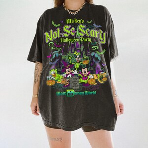 May include: A charcoal gray oversized t-shirt featuring a graphic print for Mickey's Not-So-Scary Halloween Party at Walt Disney World. The design includes Mickey, Minnie, Goofy, Donald, and Pluto in costumes, a castle, bats, and a tombstone.