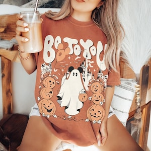 May include: A rust-coloured t-shirt with a Halloween graphic. The design includes a ghost, pumpkins, a castle, and the text "Boo To You". The graphic is in white, orange, and black.