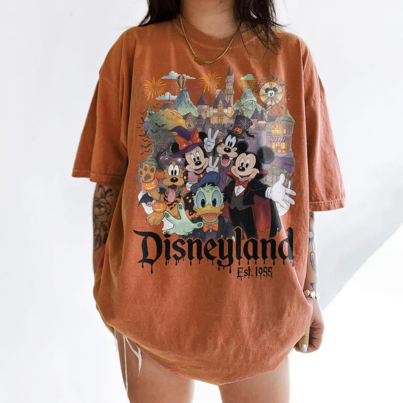 May include: An oversized, washed-out orange t-shirt featuring a graphic of Mickey Mouse, Minnie Mouse, Donald Duck, and Pluto in Halloween costumes in front of a castle. The text reads Disneyland Est. 1955 in a black, dripping, gothic-style font.