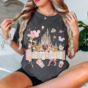 May include: Dark grey t-shirt with a colourful graphic of Disney characters and a castle. The word "Disneyworld" is displayed in a pastel font. Additional graphics include popcorn, a bow, and a Mickey Mouse balloon.