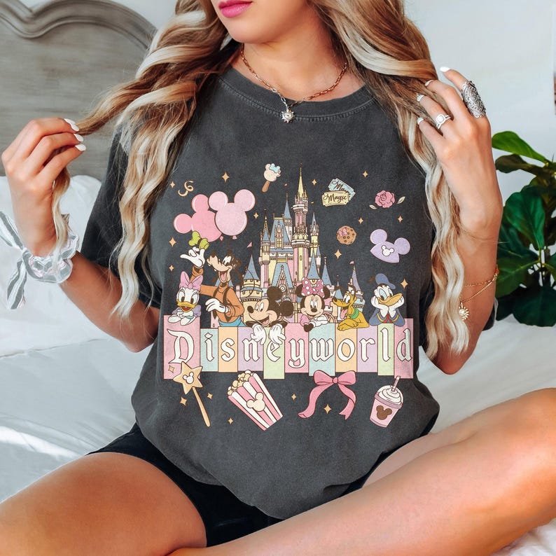 May include: Dark grey t-shirt with a colourful graphic of Disney characters and a castle. The word "Disneyworld" is displayed in a pastel font. Additional graphics include popcorn, a bow, and a Mickey Mouse balloon.