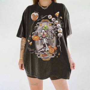 May include: Dark gray t-shirt featuring a Halloween-themed graphic. The design includes a skeleton Goofy character holding a jack-o'-lantern, a pumpkin, and a castle. The word "SPOT" is written on the shirt. Candy corn and bats are also included in the design.