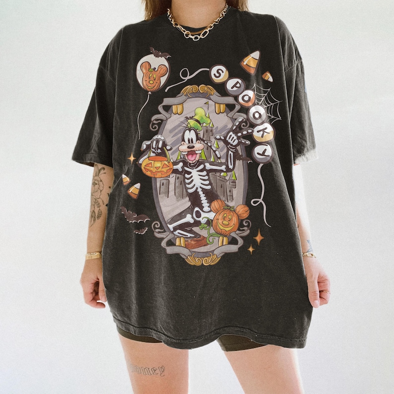 May include: Dark gray t-shirt featuring a Halloween-themed graphic. The design includes a skeleton Goofy character holding a jack-o'-lantern, a pumpkin, and a castle. The word "SPOT" is written on the shirt. Candy corn and bats are also included in the design.