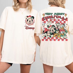 May include: Cream-coloured t-shirt with Mickey Mouse in a Santa hat and the word "Daddy". The back shows Disney characters and the text "Very Merry Christmas" above a red and white chequered pattern. Festive and seasonal.