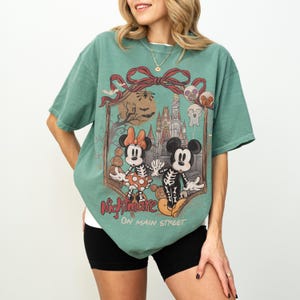 May include: Green t-shirt featuring a graphic of Mickey and Minnie Mouse as skeletons in front of a castle. The shirt has the words "Nightmare on Main Street" printed on it. The shirt is a relaxed fit.
