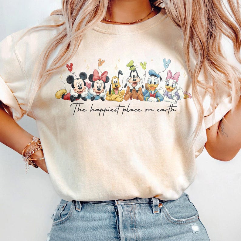 May include: Cream-colored sweatshirt featuring a line of Disney characters including Mickey Mouse, Minnie Mouse, Pluto, Goofy, Donald Duck, and Daisy Duck. The text "The happiest place on earth" is written below the characters.
