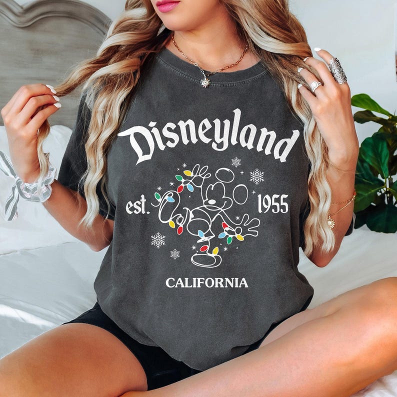 May include: Dark gray t-shirt with the text "Disneyland" in white script. Below the text is a line drawing of Mickey Mouse with colorful Christmas lights. The shirt also includes the text "est. 1955 CALIFORNIA".