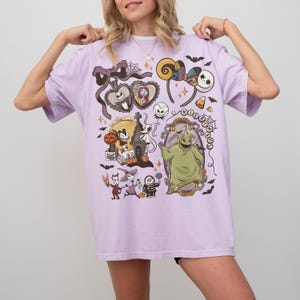 May include: A lilac t-shirt with a vibrant graphic design featuring characters and elements from "The Nightmare Before Christmas." The design includes Jack Skellington, Sally, Oogie Boogie, and other iconic imagery. The shirt has a relaxed fit.