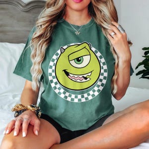 May include: Green t-shirt with a cartoon character design. The character is green with one eye, a wide smile, and small horns, set within a white circle with a black and white chequered border. The shirt is a solid green colour.