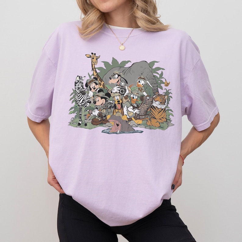 K&ouml;nnte beinhalten: Helllila T-Shirt mit einer Grafik von Zeichentrickfiguren in Safari-Outfits. Das Design zeigt ein Zebra, eine Giraffe, einen Elefanten, einen Tiger und andere Charaktere. Das T-Shirt hat einen Rundhalsausschnitt.