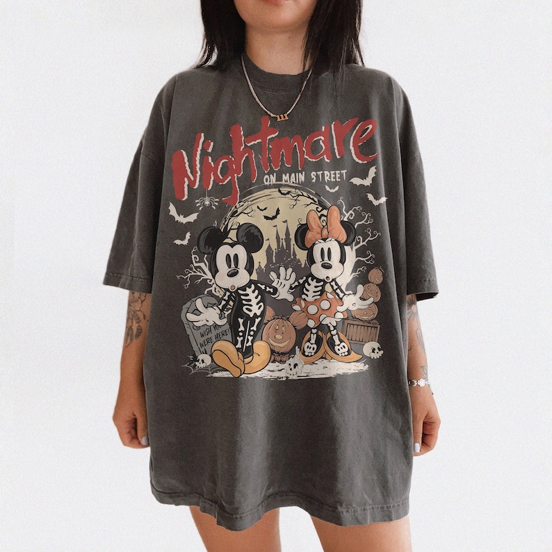 May include: Dark gray t-shirt featuring a Halloween-themed graphic. The design includes Mickey and Minnie Mouse dressed as skeletons, the words "Nightmare on Main Street," bats, and a full moon. The shirt has a vintage, oversized fit.