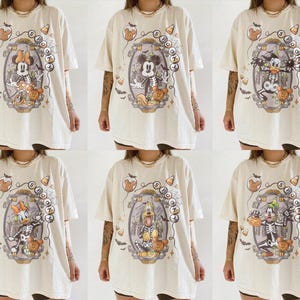 May include: Cream-colored Halloween t-shirts featuring various Disney characters as skeletons. Each shirt has a unique design within an oval frame, with candy corn and spooky accents. The shirts are oversized and have short sleeves.