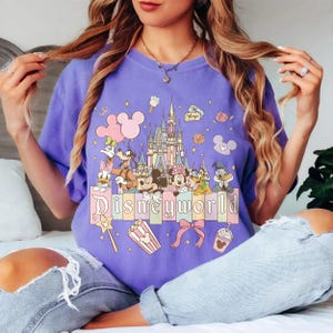 May include: A lilac t-shirt with a Disney World graphic. The design features Mickey and Minnie Mouse, Donald Duck, Goofy, and a castle, with the word "Disneyworld" across the bottom in a pastel font.