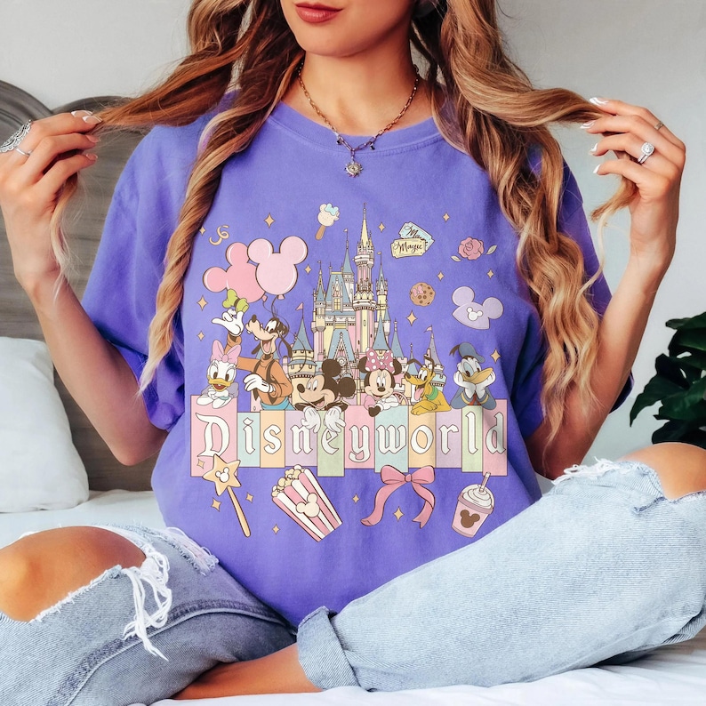May include: A lilac t-shirt with a Disney World graphic. The design features Mickey and Minnie Mouse, Donald Duck, Goofy, and a castle, with the word "Disneyworld" across the bottom in a pastel font.