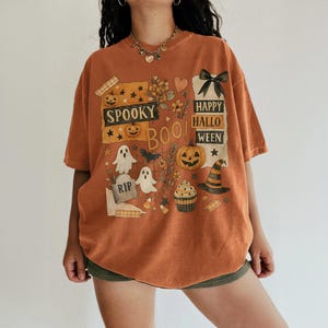 K&ouml;nnte beinhalten: Orangefarbenes T-Shirt mit einem Halloween-Motiv. Das Design umfasst K&uuml;rbisse, Geister, einen Hexenhut und die Worte "Spooky Boo! Happy Halloween". Das kurz&auml;rmelige T-Shirt hat eine lockere Passform, ideal f&uuml;r den Herbst.