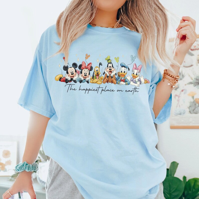 May include: Light blue t-shirt featuring a graphic of Disney characters including Mickey, Minnie, Goofy, Donald, and Daisy Duck. The text "The happiest place on earth" is printed below the characters. The shirt has short sleeves.