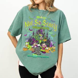May include: A sage green short-sleeve t-shirt featuring a graphic of Disney characters in Halloween costumes. The text reads Mickey's Not-So-Scary Halloween Party and Walt Disney World. The design includes a castle, bats, and pumpkins.