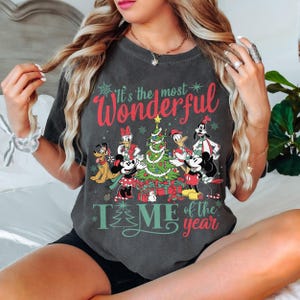 May include: Dark gray t-shirt featuring a festive design with Disney characters around a Christmas tree. The text reads "It's the most Wonderful TIME of the year" in red and green. The shirt has a vintage, worn look.