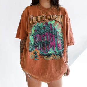 May include: Burnt orange t-shirt featuring a graphic of a haunted mansion with the text "The Haunted Mansion". The design includes a spooky house, ghosts, pumpkins, and a cartoon character. The shirt has short sleeves.