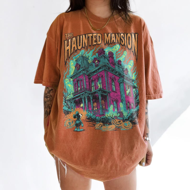 May include: Burnt orange t-shirt featuring a graphic of a haunted mansion with the text "The Haunted Mansion". The design includes a spooky house, ghosts, pumpkins, and a cartoon character. The shirt has short sleeves.