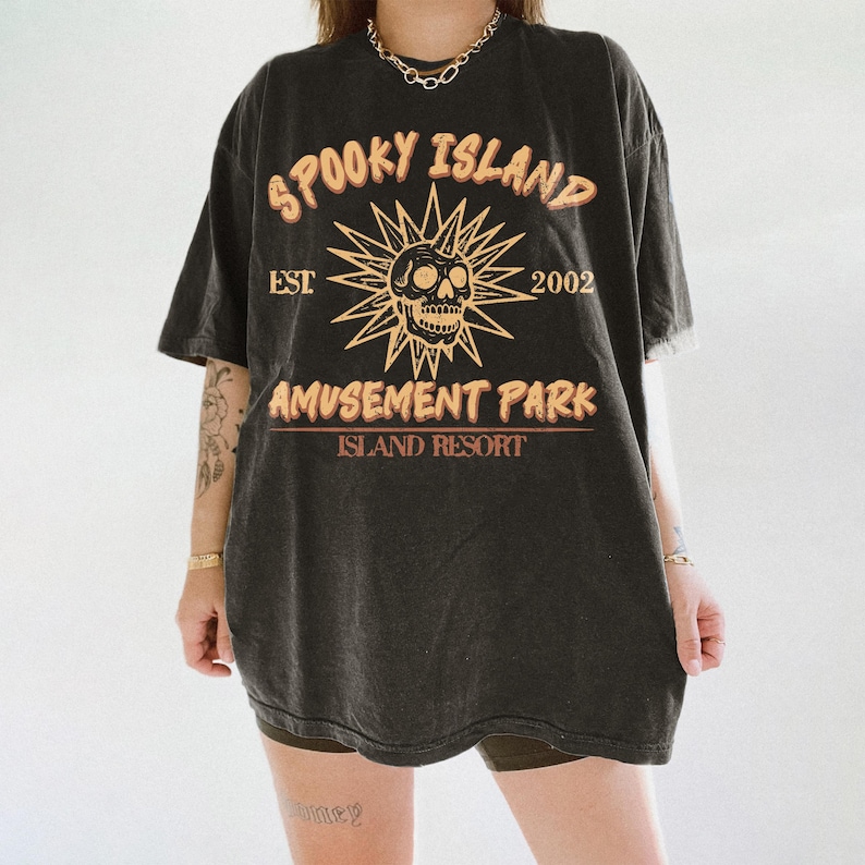 May include: Black t-shirt with the text "SPOOKY ISLAND AMUSEMENT PARK ISLAND RESORT" in orange and yellow. The design includes a skull with sun rays and the year "2002". The shirt has a vintage, worn look.