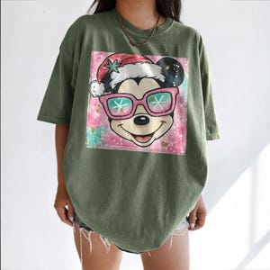 K&ouml;nnte beinhalten: Olivgr&uuml;nes T-Shirt mit einem Aufdruck von Mickey Mouse mit Sonnenbrille und Weihnachtsm&uuml;tze. Die Grafik hat einen rosa-t&uuml;rkisfarbenen Aquarellhintergrund mit Schneeflocken-Details. Das Shirt hat kurze &Auml;rmel und eine lockere Passform.