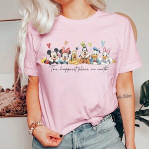 May include: A light pink t-shirt featuring a graphic of Disney characters including Mickey, Minnie, Goofy, Donald, and Daisy Duck. The text "The happiest place on earth" is printed below the characters. The shirt has short sleeves.