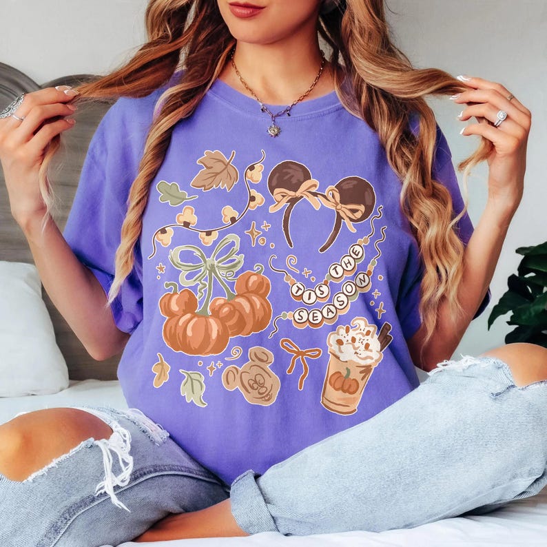 May include: A purple short-sleeve t-shirt featuring a graphic print of orange pumpkins, brown mouse ears with bows, a string of lights, and a cup of coffee with the text 'TIS THE SEASON' written on a string of beads in a decorative fall theme.