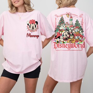 May include: Light pink t-shirt featuring a Minnie Mouse graphic with the word "Mommy" on the front. The back of the shirt displays a Disney World design with Mickey, Minnie, Goofy, Donald, and Pluto. The design includes Christmas trees and the text "Disneyworld 1971".