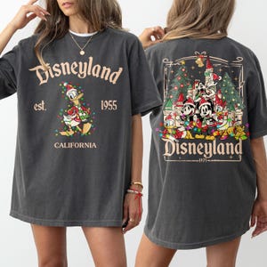May include: Dark gray t-shirt with Disneyland graphics. The front features Donald Duck in a Santa hat and Christmas lights, with "Disneyland est. 1955 California" text. The back displays Disney characters in front of a castle and Christmas trees.