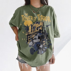 May include: Olive green t-shirt featuring a graphic of Mickey Mouse in a bellhop uniform, with the words "The Tower of Terror" and "Hollywood Tower" in yellow and white. The shirt has a vintage, distressed look.