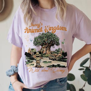 May include: A light purple t-shirt featuring the text "Disney's Animal Kingdom" in orange script above an illustration of the Tree of Life and Mickey Mouse. The shirt also has the text "Wild about Disney" in a cursive font.