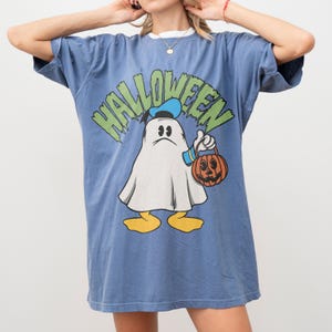 May include: Blue t-shirt with a Halloween graphic featuring a cartoon ghost holding a jack-o'-lantern. The word "HALLOWEEN" is printed above the ghost in green and yellow letters. The shirt has a white collar.