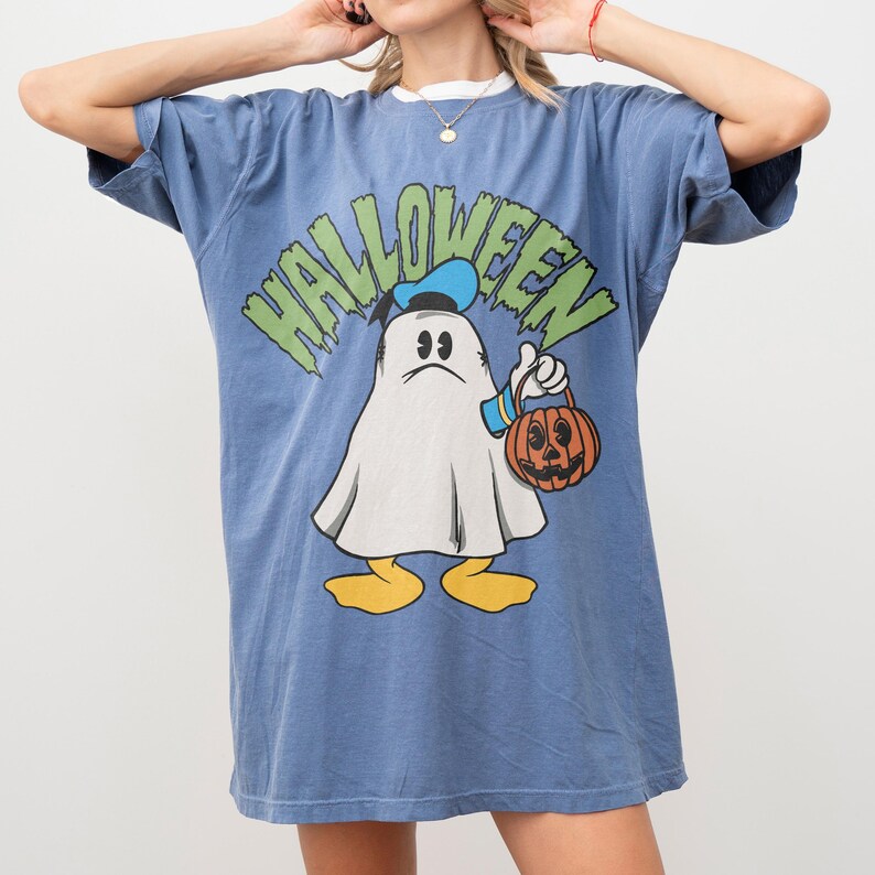 May include: Blue t-shirt with a Halloween graphic featuring a cartoon ghost holding a jack-o'-lantern. The word "HALLOWEEN" is printed above the ghost in green and yellow letters. The shirt has a white collar.