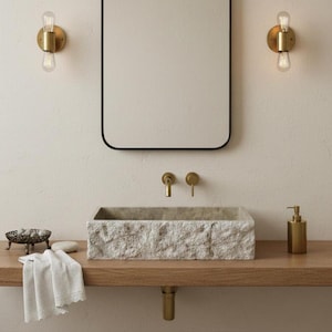May include: A bathroom vanity with a rectangular stone sink, a wooden countertop, and a black-framed mirror. Two brass sconces with Edison bulbs flank the mirror. A brass faucet and soap dispenser complete the minimalist design.