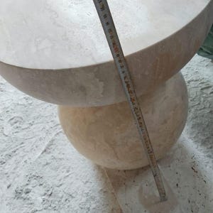 May include: A light beige stone table with a rounded top and a spherical base. The table is being measured with a ruler. The top of the table appears to be approximately 24 inches in diameter. The table is sitting on a dusty surface.