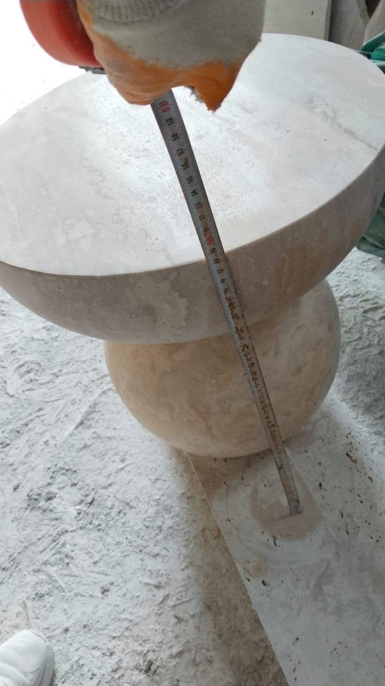 May include: A light beige stone table with a rounded top and a spherical base. The table is being measured with a ruler. The top of the table appears to be approximately 24 inches in diameter. The table is sitting on a dusty surface.