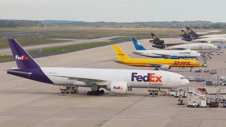 May include: A fleet of cargo airplanes parked on a tarmac. The lead plane is white with a purple tail and the FedEx logo. Other planes include DHL, UPS, and Star Air. Ground support equipment is visible.