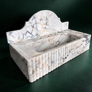 Puede incluir: Un lavabo rectangular de mármol con respaldo festoneado y laterales acanalados. El lavabo cuenta con una pila y un estante con tres agujeros. El mármol tiene una base blanca con vetas grises y doradas.