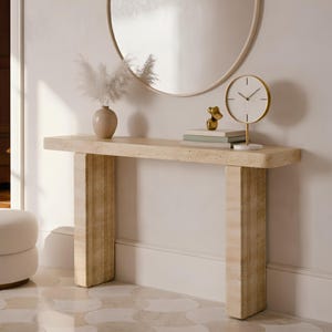 Può includere: Una consolle in pietra beige con due gambe rettangolari. Uno specchio rotondo è appeso sopra il tavolo. Un vaso con fiori secchi, un orologio dorato e libri sono sul tavolo. A sinistra c'è un piccolo pouf rotondo imbottito.