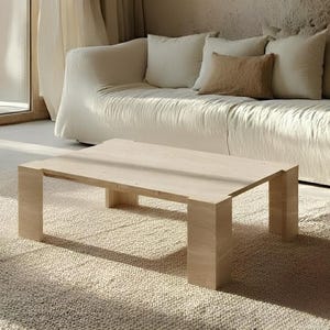 May include: A light-colored rectangular coffee table with a minimalist design. The table has a solid, blocky construction with four thick legs. It sits on a textured, light-colored rug, with a white sofa and pillows in the background.